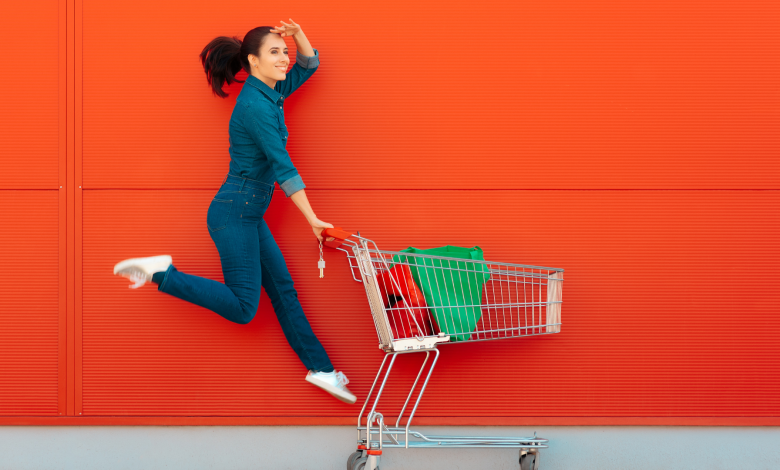 #PraCegoVer: Uma imagem com fundo cor vermelho e uma mulher pulando e ao mesmo tempo empurando um carinho de compras de supermercado